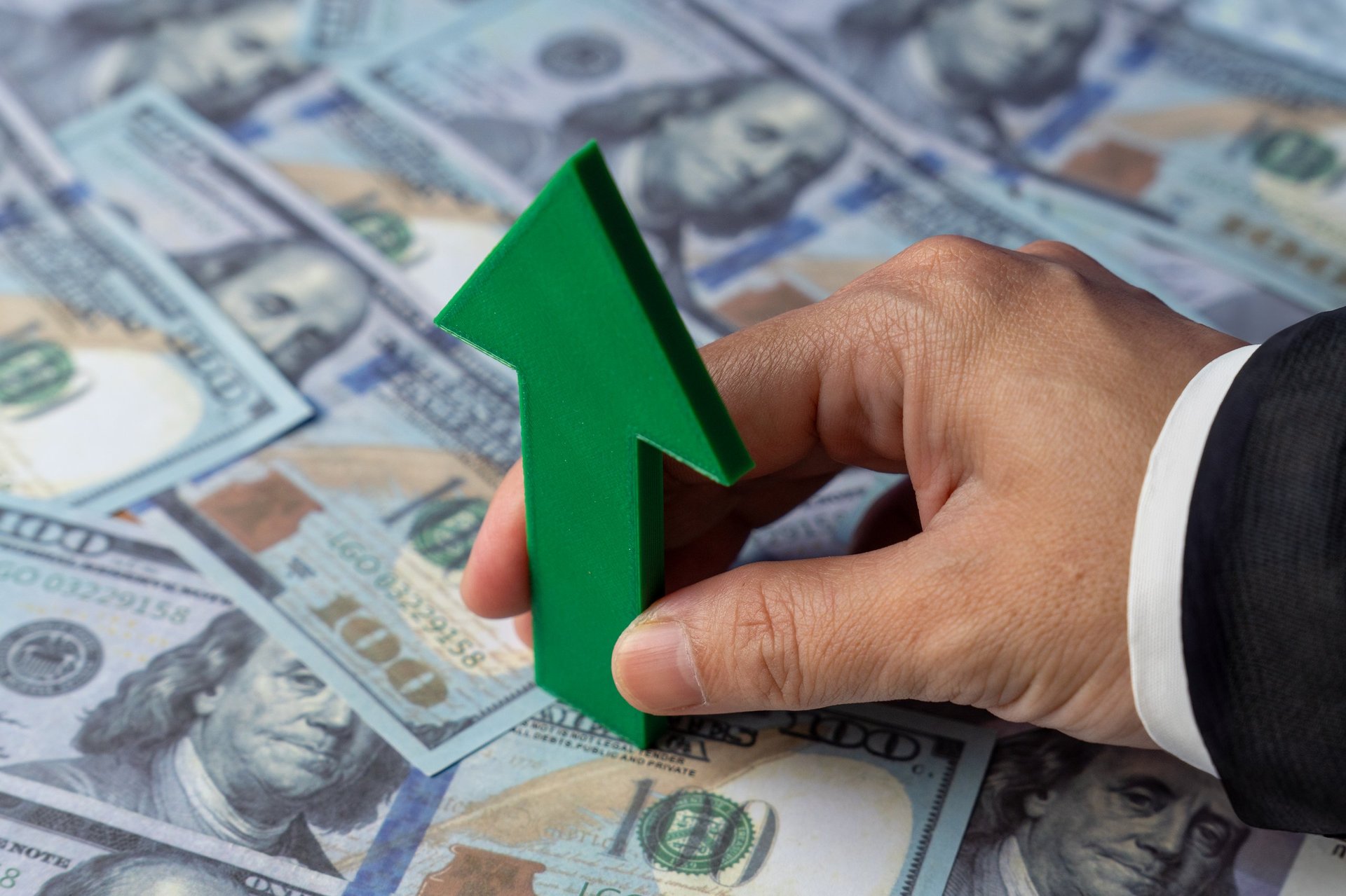 A hand holds a green upward-pointing arrow over a spread of US one-hundred dollar bills